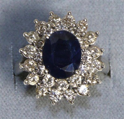 Lot 723 - A 14 Carat White Gold Sapphire and Diamond Cluster Ring, an oval cut sapphire in a claw...