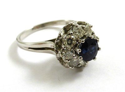 Lot 2151 - An 18ct White Gold Sapphire and Diamond Cluster Ring, 1975, a round cut sapphire above a border...