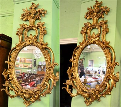 Lot 1411 - A pair of gilt two-branch girandoles, modern, with oval mirror plates and scrolled frames...