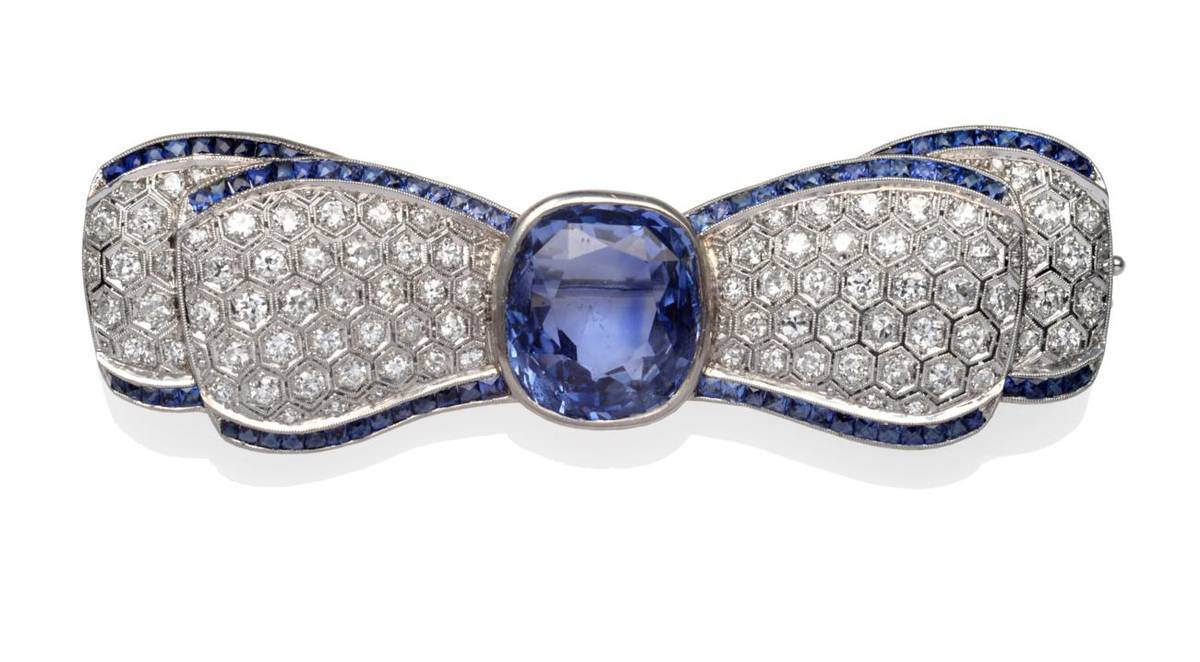 Lot 372 - A Sapphire and Diamond Bow Brooch, the cushion mixed cut sapphire in a white rubbed over...