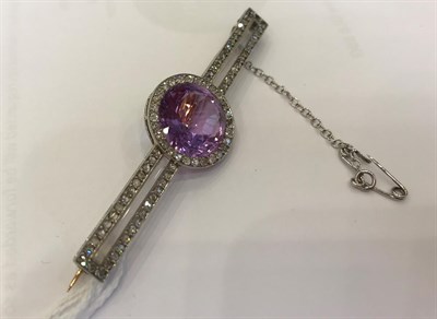 Lot 1045 - A Diamond and Pinkish-Purple Stone Bar Brooch, the round cut pinkish-purple stone within a...