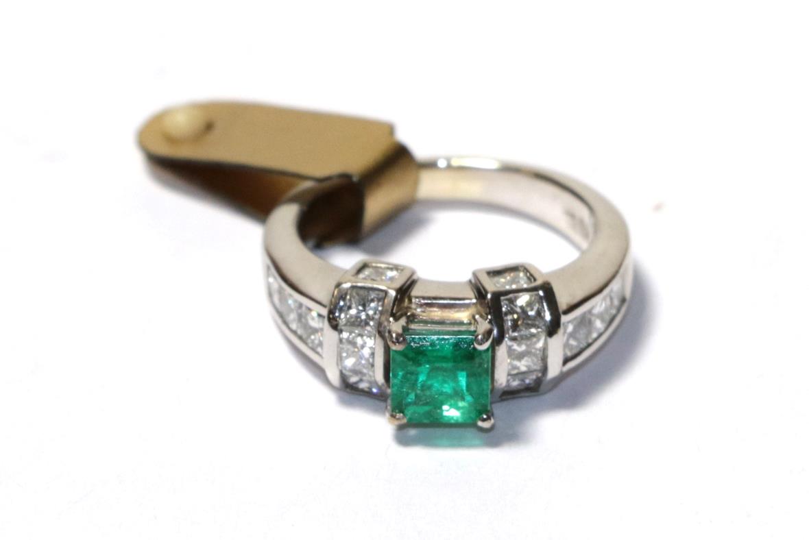 Lot 97 - An emerald and diamond ring, the emerald-cut emerald in a white four claw setting, flanked by a...