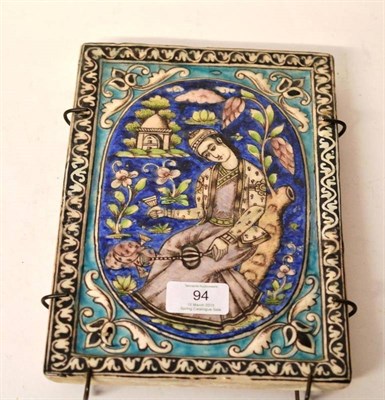 Lot 94 - A Qajar Pottery Tile, 19th century, painted with a seated gentleman holding a glass and decanter in