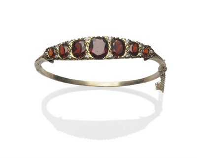 Lot 359 - A Garnet and Diamond Set Bangle, seven graduated oval cut garnets spaced by pairs of brilliant...